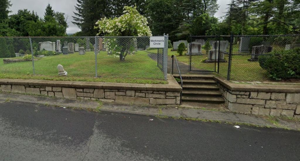 Workmen's Circle Cemetery viewed from Stillson Road, rows of gravestones on green lawn behind ornate iron fence